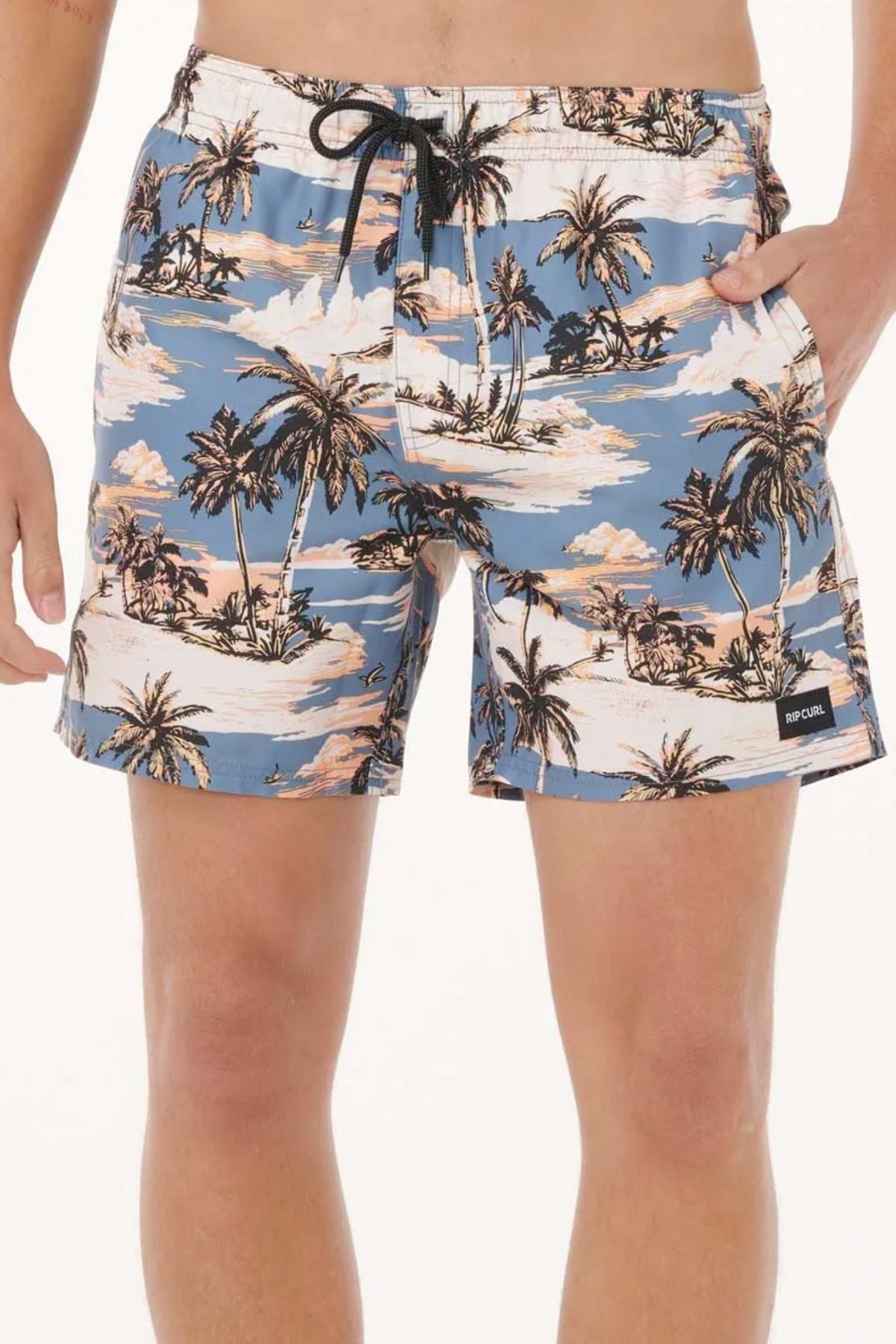 Someone wears Rip Curl Dreamers Volley shorts in blue, featuring a tropical palm tree and beach print, black drawstring waist, and a black logo patch on the left leg. These quick-dry shorts provide comfort and style for beach days.