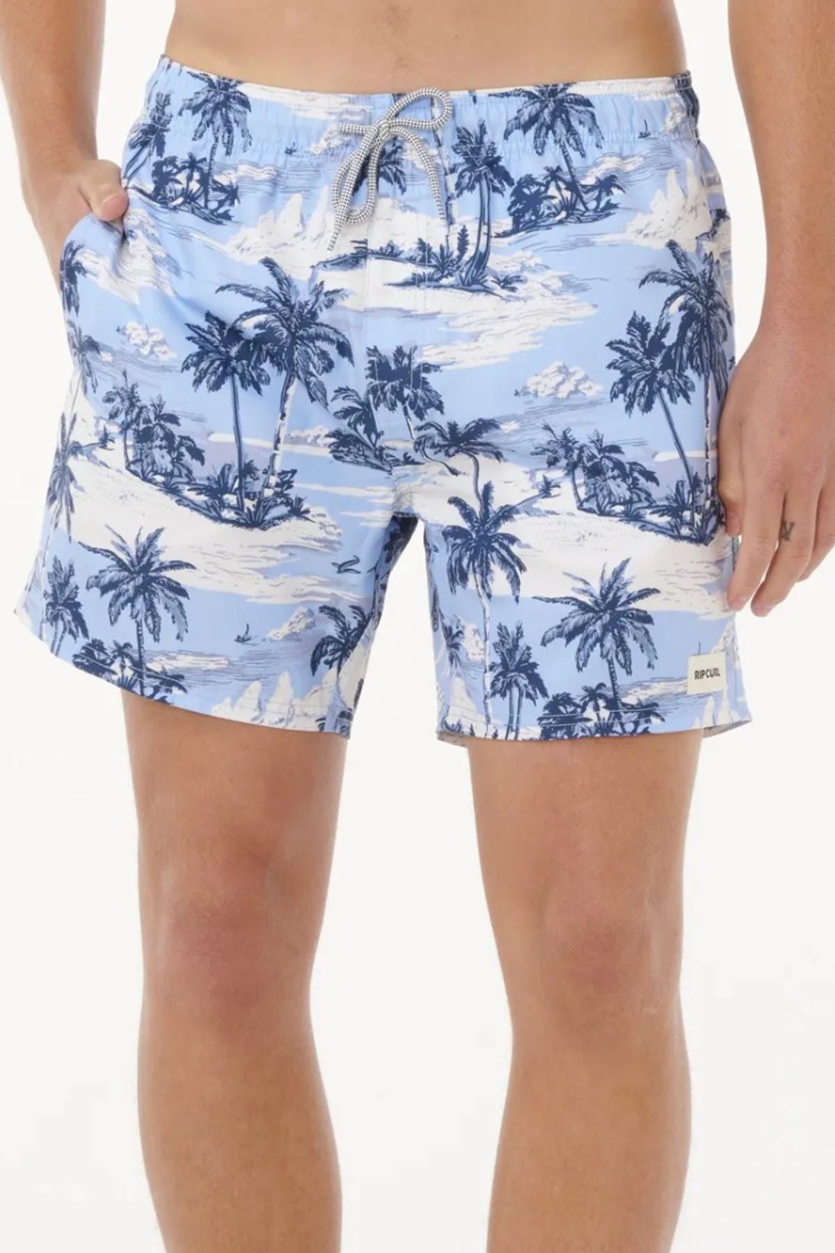 Wearing the Rip Curl Dreamers Volley shorts in light blue with a white and navy tropical island and palm tree print, a person stands against a plain backdrop with their left hand in their pocket, highlighting this stylish, sustainable swimwear.