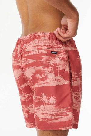 A person wearing the Rip Curl Dreamers Volley, which are stylish red tropical-patterned board shorts. These shorts, crafted from recycled fabric, showcase an island and palm tree design along with a small back pocket and a logo patch. The individual stands with one hand adjusting the waistband against a plain white background.