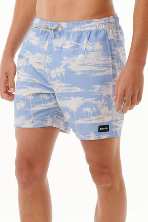 A person wearing the Rip Curl Dreamers Volley, a pair of stylish light blue board shorts adorned with a white tropical palm tree pattern. These shorts are crafted from recycled rip suede fabric and feature a black label with text on the front. The person's hand rests by their side against a plain white background.