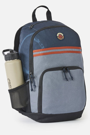 A navy and grey Rip Curl Evo 24L Rinsed Backpack with a mesh side pocket holding a beige water bottle.