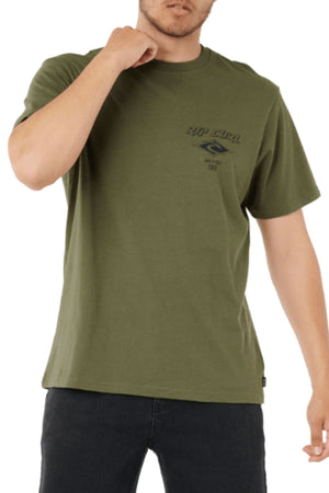 Someone wearing the Rip Curl Fade Out Icon Tee in olive green, featuring a small dark logo and text on the left chest, paired with dark pants. The relaxed fit t-shirt gives a casual vibe; the person's face is partially out of frame.