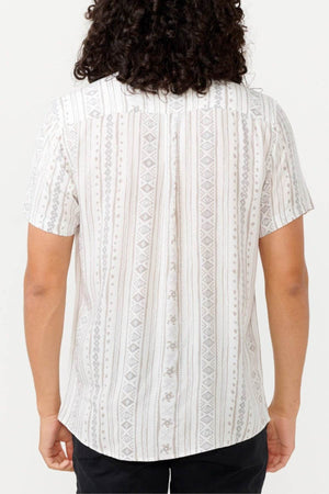 Rear view of a person wearing a short-sleeve, light-colored Rip Curl Fun Times Short Sleeve Shirt featuring a vertical geometric pattern. Made from eco-friendly fabric, their viscose top pairs well with long, curly hair and dark pants. The background is plain white.