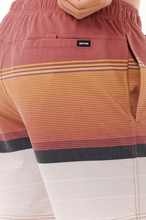 A person is wearing the Rip Curl Fun Times Volley shorts, adorned with red and orange stripes. The design includes a white section towards the hem and a back pocket branded with "Rip Curl." Equipped with quick-dry technology, these shorts are perfect for summer escapades.