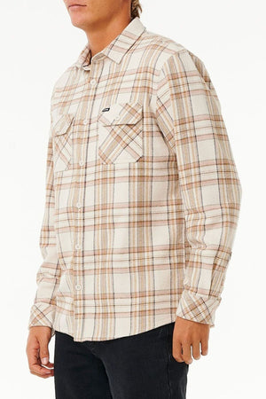 A person in the Rip Curl Griffin Flannel Shirt (beige and brown plaid cotton flannel) with black pants stands before a plain white background, their face partially out of frame.
