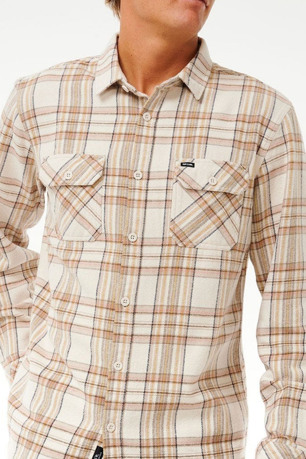 Someone stands against a plain light background wearing the Rip Curl Griffin Flannel Shirt, featuring beige, brown, and white plaid with two chest pockets. Their face is partially out of frame.