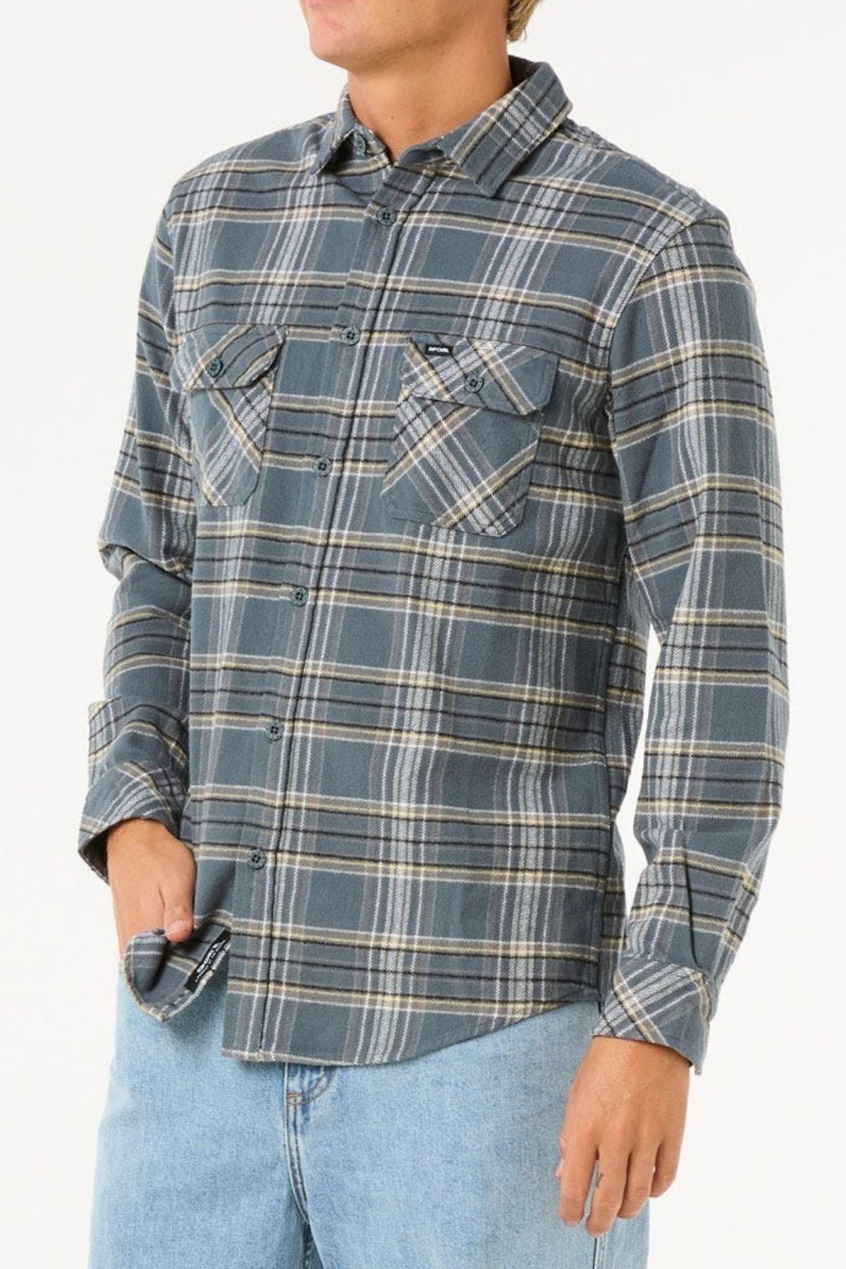 A person wearing a gray and blue plaid Rip Curl Griffin Flannel Shirt with buttoned chest pockets and light blue jeans stands with one hand in their pocket against a plain white background.