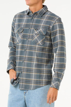 A person wearing a gray and blue plaid Rip Curl Griffin Flannel Shirt with buttoned chest pockets and light blue jeans stands with one hand in their pocket against a plain white background.