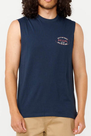 An individual is dressed in a Rip Curl Hallmark Muscle sleeveless t-shirt in navy blue, featuring the logo prominently on the chest, paired with beige pants. The muscle fit design offers a snug appearance, complemented by the use of responsibly sourced materials. The face is not shown.