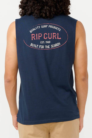 A person is featured standing with their back turned, wearing the Rip Curl Hallmark Muscle sleeveless navy shirt adorned with the phrase "Built for the Search" on its back. Complemented by beige pants, this garment honors Rip Curl's surfing legacy since 1969 and is crafted from responsibly sourced materials.