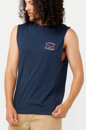 A man with curly hair wears the Rip Curl Hallmark Muscle navy blue sleeveless t-shirt, showcasing its muscle fit with the logo prominently displayed on the chest. He looks to the side, paired with beige pants against a plain white background.