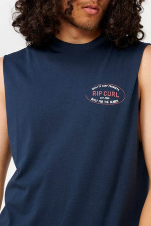 A person with curly hair is wearing the Rip Curl Hallmark Muscle shirt, a navy blue sleeveless design in muscle fit, featuring the "Rip Curl" logo on the chest. The emblem includes the phrase "Quality Surf Products, Est. 1969, Built for the Search," perfectly capturing the ethos of this product.