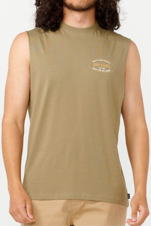 A person with long dark hair is donning the Rip Curl Hallmark Muscle shirt in olive green, featuring a sleeveless design that highlights its muscle fit. The small "RIP CURL" logo embellishes the left side, and paired with beige pants, this ensemble projects a laid-back vibe against the simple light background.