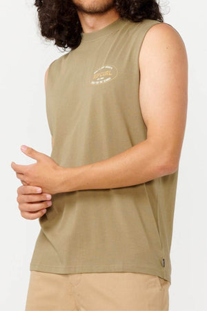 A person with long hair is wearing the "Rip Curl Hallmark Muscle," a sleeveless olive green shirt in a muscle fit, crafted from 100% cotton. Complemented by beige pants and arms crossed at waist height, they exude confidence against a plain white background.