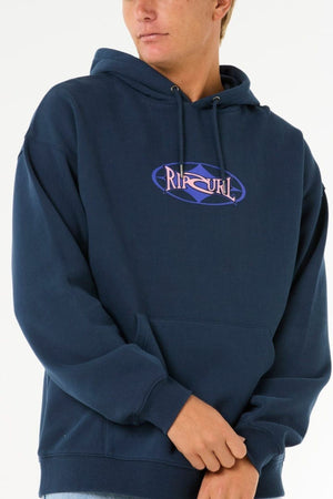 A person in a dark blue Rip Curl Heritage Oval Hood, made from soft knit fabric with a front pocket and purple chest logo, stands against a plain white background.