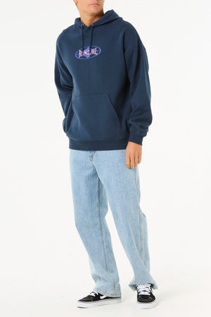 A person wears a navy blue Rip Curl Heritage Oval Hood with a purple logo, paired with light blue jeans and black-and-white sneakers, standing against a plain white background.