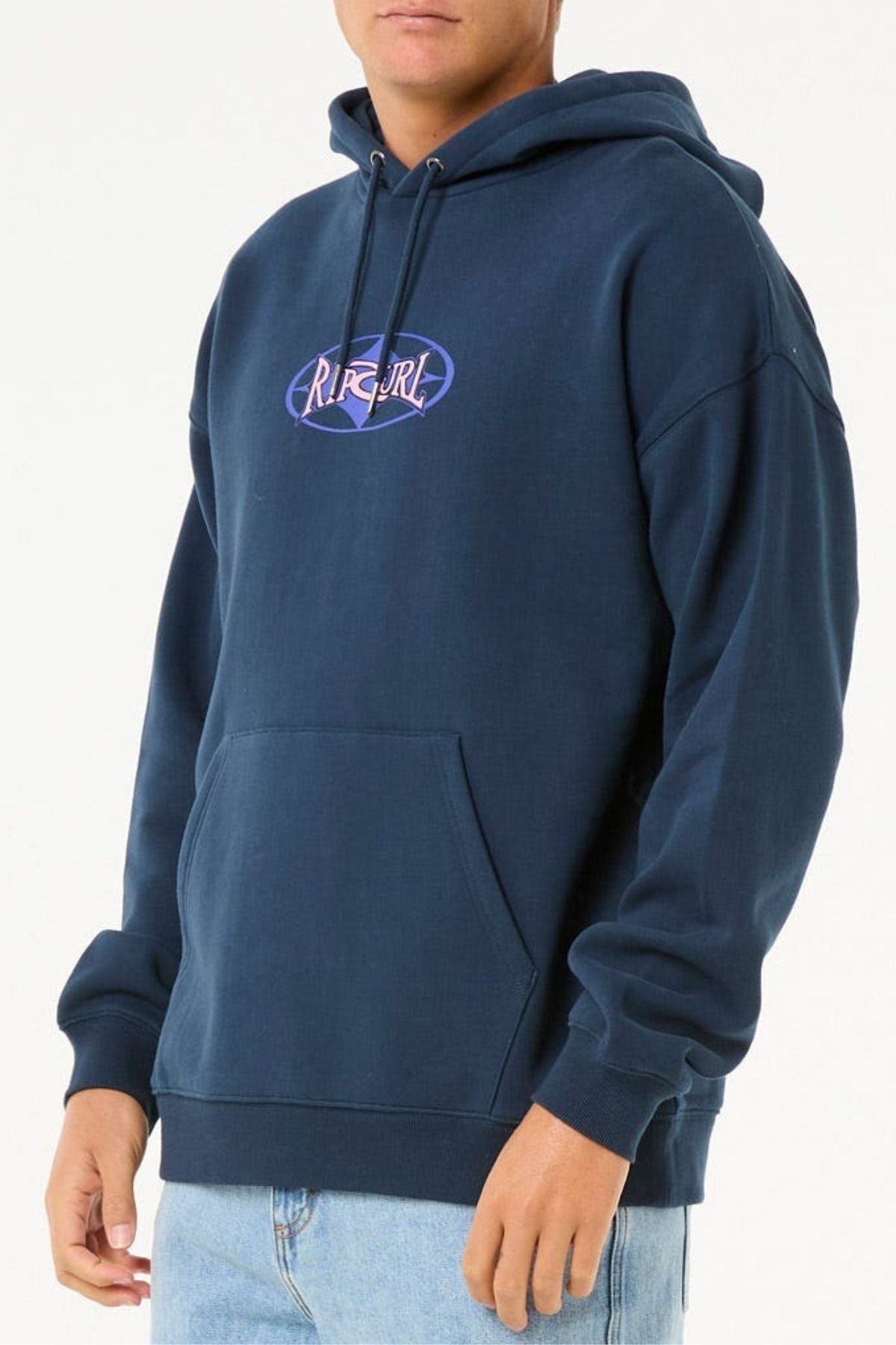 A person wears a dark blue Rip Curl Heritage Oval Hood, made from soft knit material with a purple “RIP CURL” logo and front pocket, paired with light blue jeans against a plain white background.