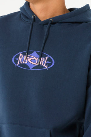 Person in a blue Rip Curl Heritage Oval Hood, made from soft knit fabric. The hoodie displays the Rip Curl logo in purple and pink within an oval design. Only the upper torso is shown.