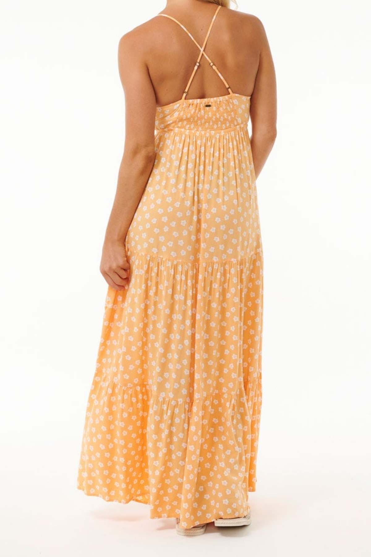 The individual is dressed in a long, sleeveless orange dress adorned with white floral designs, made from soft Viscose. This outfit includes a shirred back and thin straps crossing over the shoulders, echoing the style of the Rip Curl High Tide Ditsy Maxi Skirt. They are posing with their back to the camera against a plain backdrop.