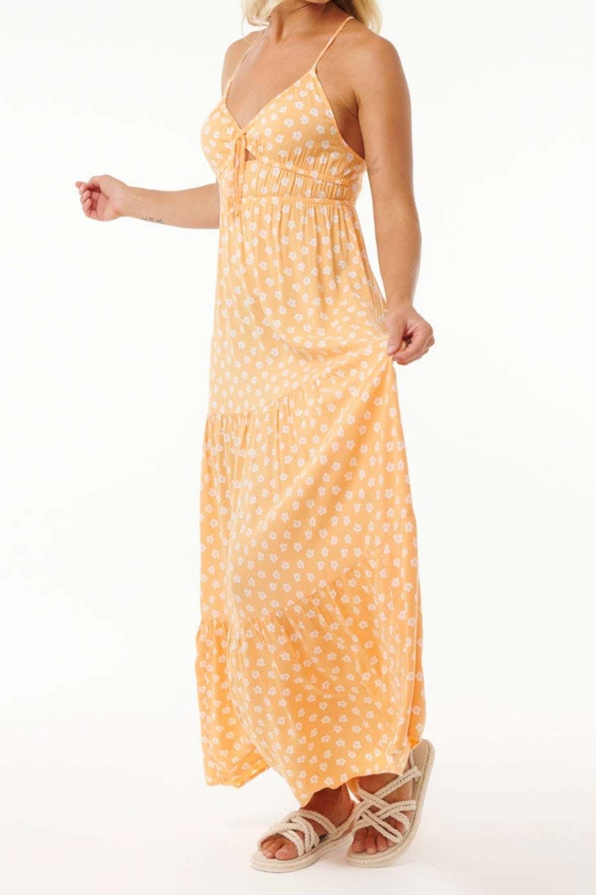 A person is wearing the Rip Curl High Tide Ditsy Maxi Skirt, an orange skirt with white floral patterns made of Lenzing Ecovero Poplin. The skirt is long and flowy, and they are standing while holding it with one hand, paired with white sandals.