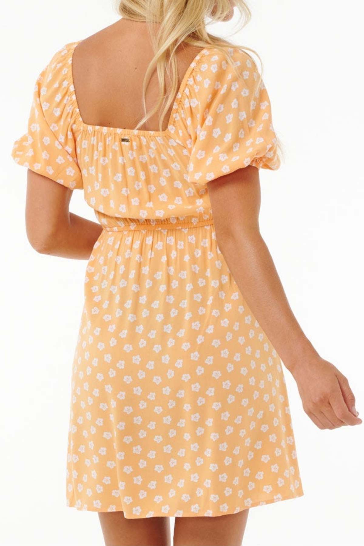 The sentence could be rewritten as: "From the back, a person showcases the elegance of the Rip Curl High Tide Ditsy Mini Dress, distinguished by its yellow fabric adorned with a white floral pattern. With its square neckline and short puff sleeves, this dress enhances any trendy wardrobe and beautifully accents long, wavy blonde hair.