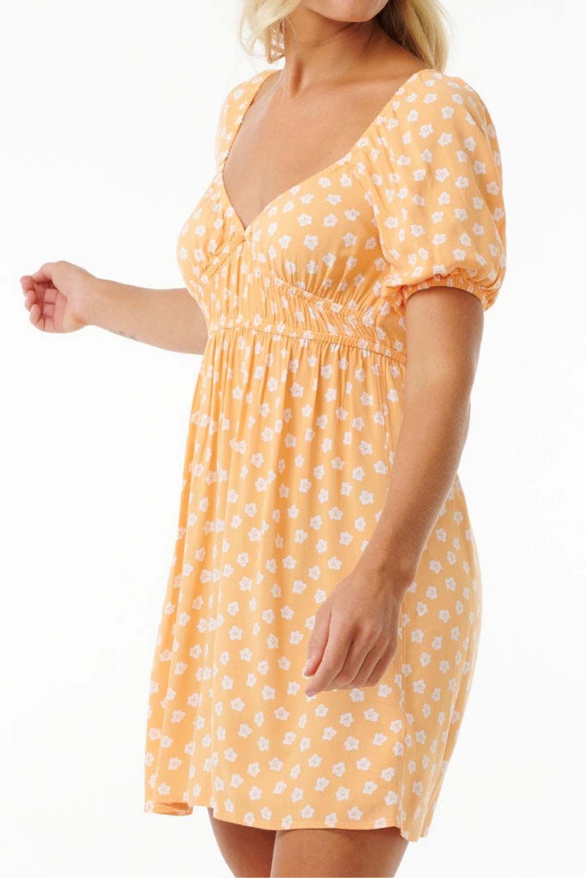 A person poses against a simple backdrop, dressed in a Rip Curl High Tide Ditsy Mini Dress featuring an orange hue with white floral designs. This dress's short sleeves and cinched waist create a relaxed, summery feel ideal for a trendy wardrobe. The individual's head isn't visible in the image.
