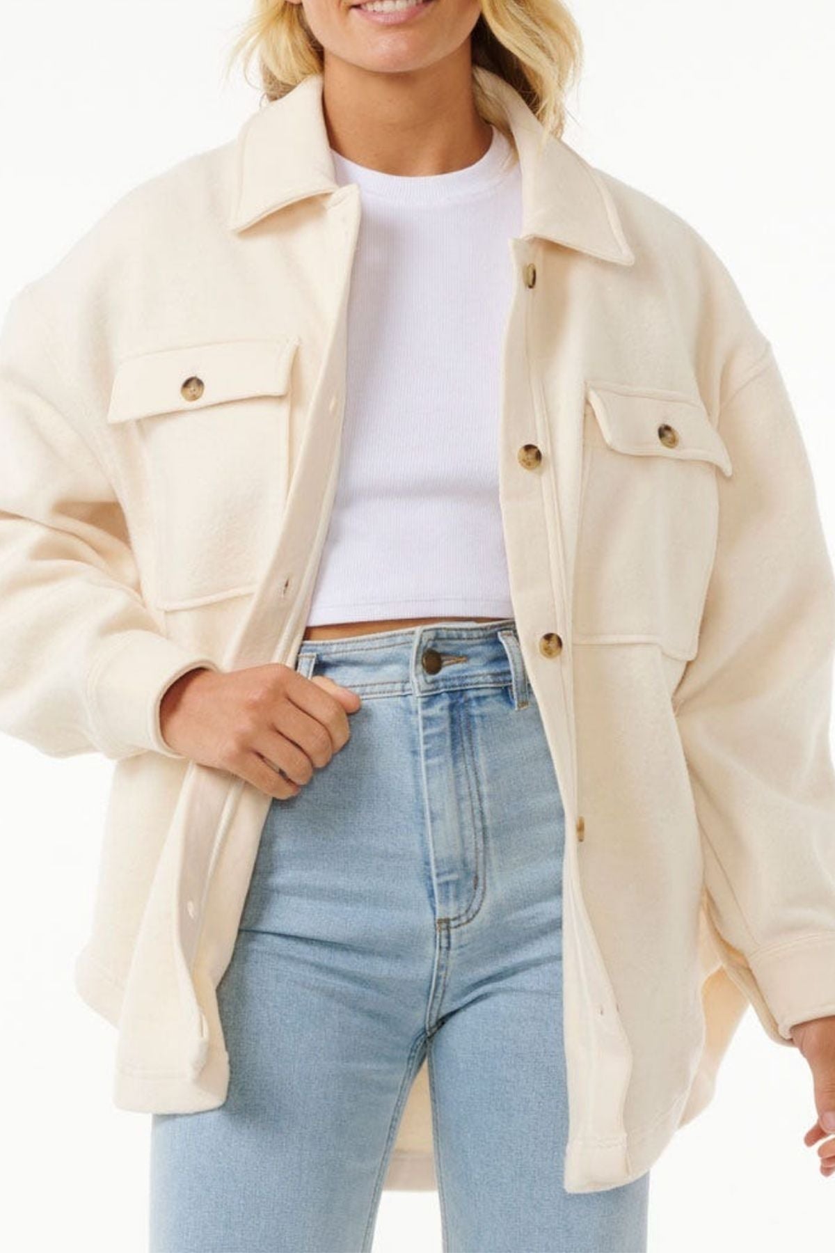 A woman wears the Rip Curl High Tide Fleece Shacket over a white cropped top and light blue high-waisted jeans, pairing comfort and style for adventurous outings, set against a crisp white background.