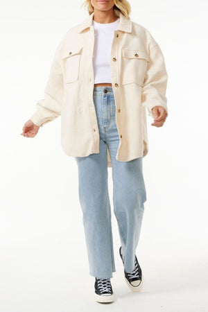 A person in a cream Rip Curl High Tide Fleece Shacket over a white top, light blue high-waisted jeans, and black-and-white sneakers stands against a white background. Their face is partially out of frame—perfect for adventurous styles.