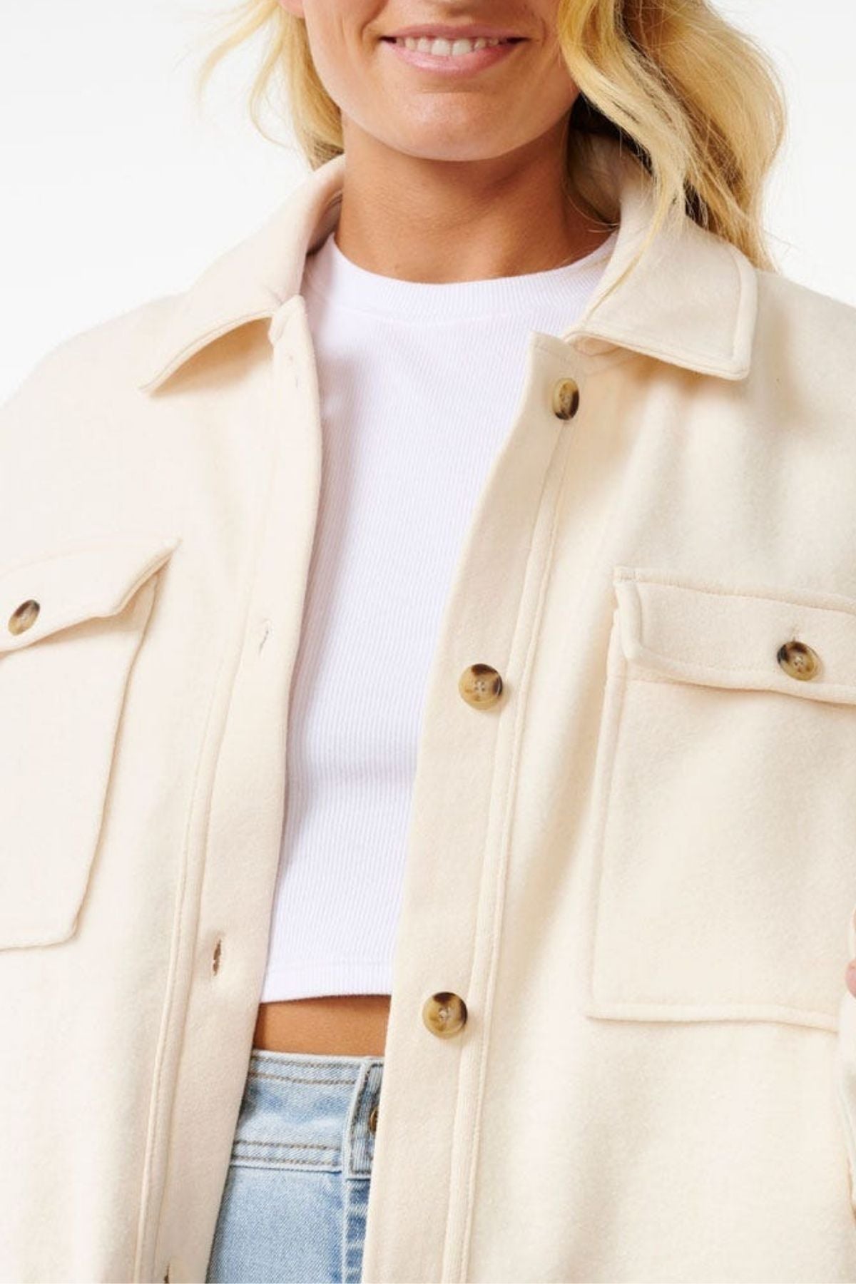 A woman with blonde hair, smiling slightly, wears a cream Rip Curl High Tide Fleece Shacket with large pockets over a white ribbed crop top and light blue jeans—perfect for adventurous individuals.