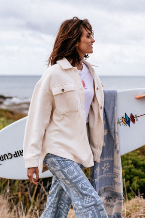 A woman with shoulder-length hair smiles outdoors near the ocean, carrying a surfboard under her arm while wearing the Rip Curl High Tide Fleece Shacket and patterned blue pants by grassy coastal scenery.