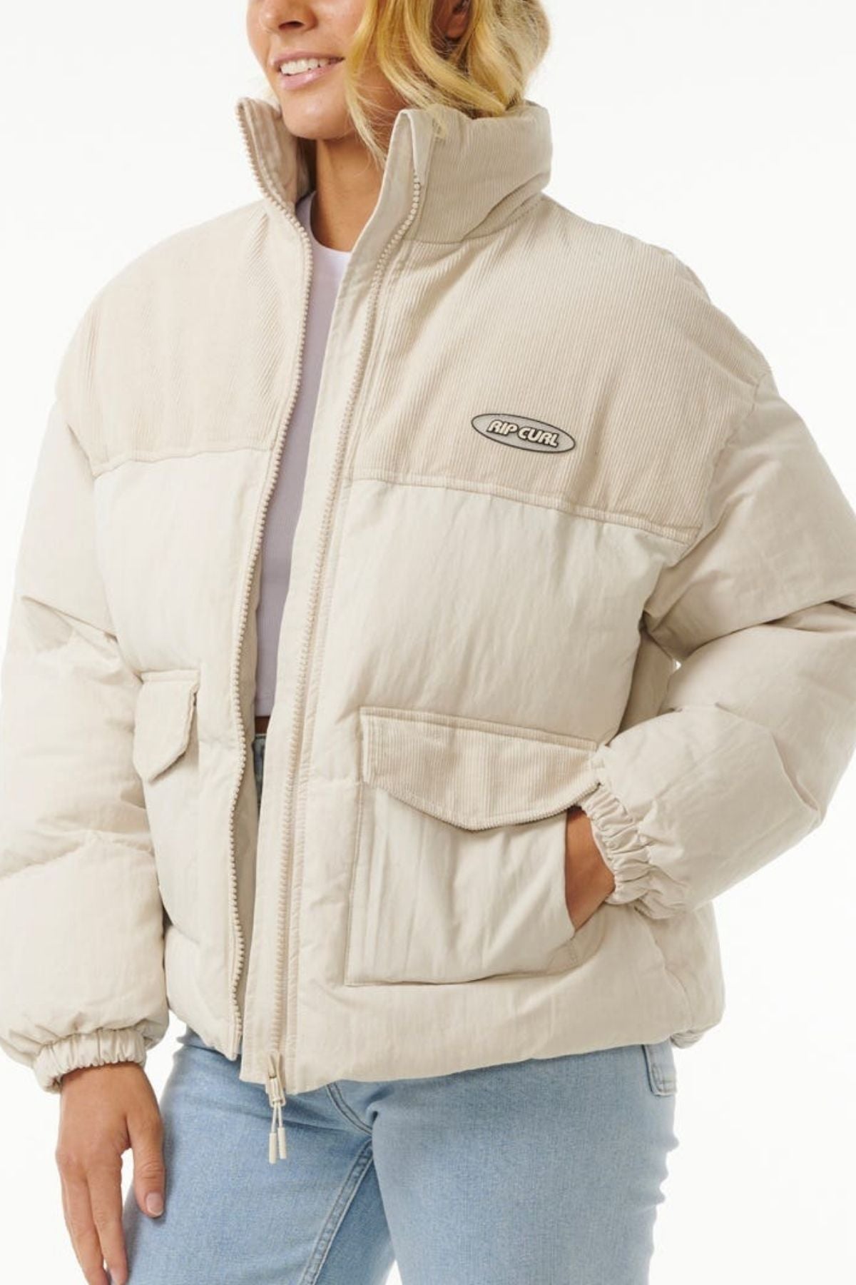 A person wears the Rip Curl High Tide Mixed Cord Puffer, a light beige jacket with a high collar, large front pockets, an embroidered chest logo, and cotton cord panels on the collar and shoulders, paired with light blue jeans.