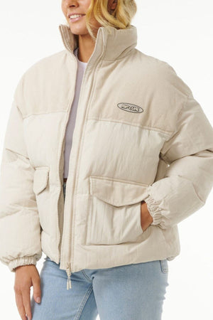 A person wears the Rip Curl High Tide Mixed Cord Puffer, a light beige jacket with a high collar, large front pockets, an embroidered chest logo, and cotton cord panels on the collar and shoulders, paired with light blue jeans.