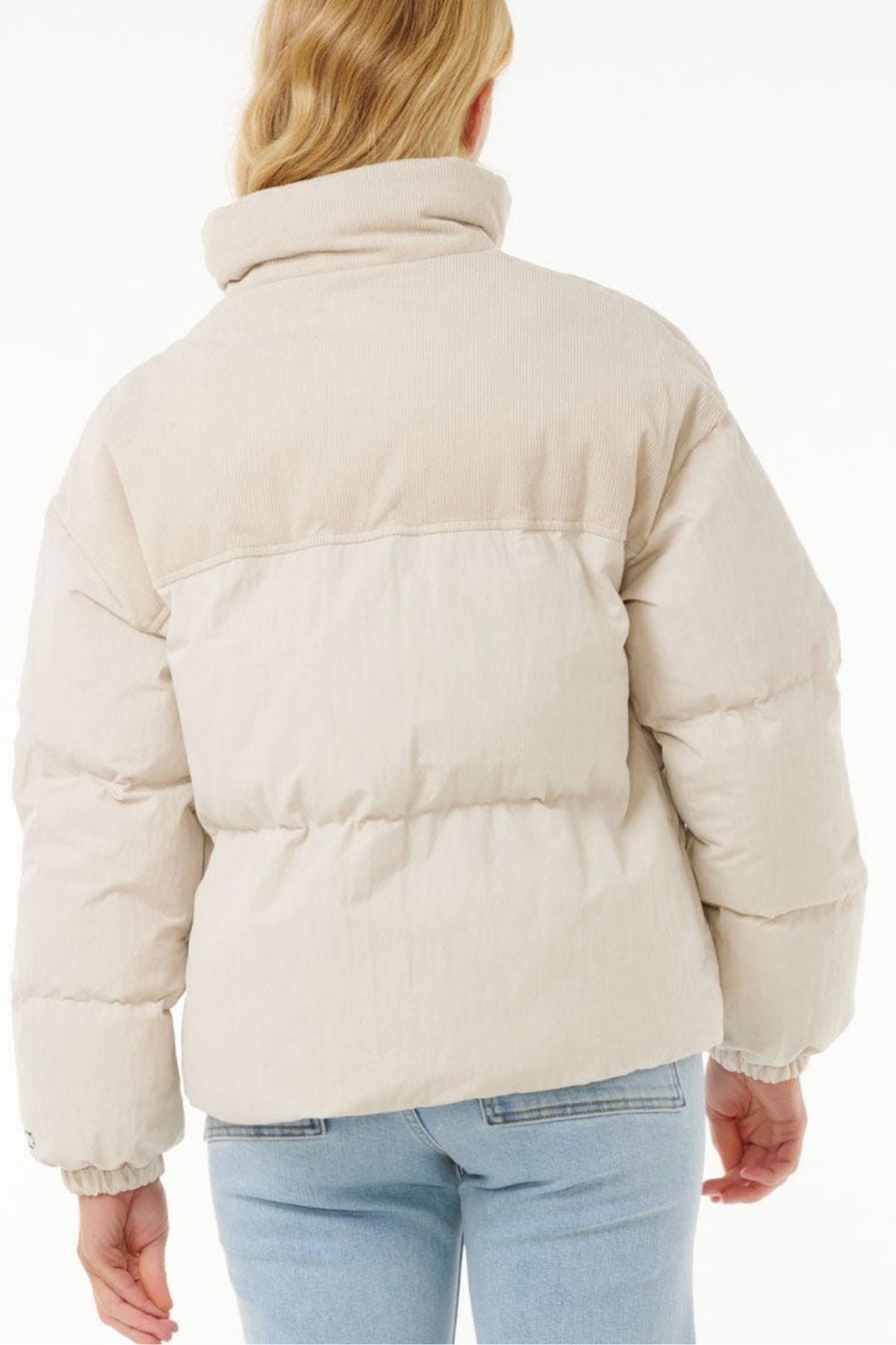 A person with long, blonde hair is shown from the back against a plain white background, wearing a light beige Rip Curl High Tide Mixed Cord Puffer with an embroidered logo and light blue jeans.