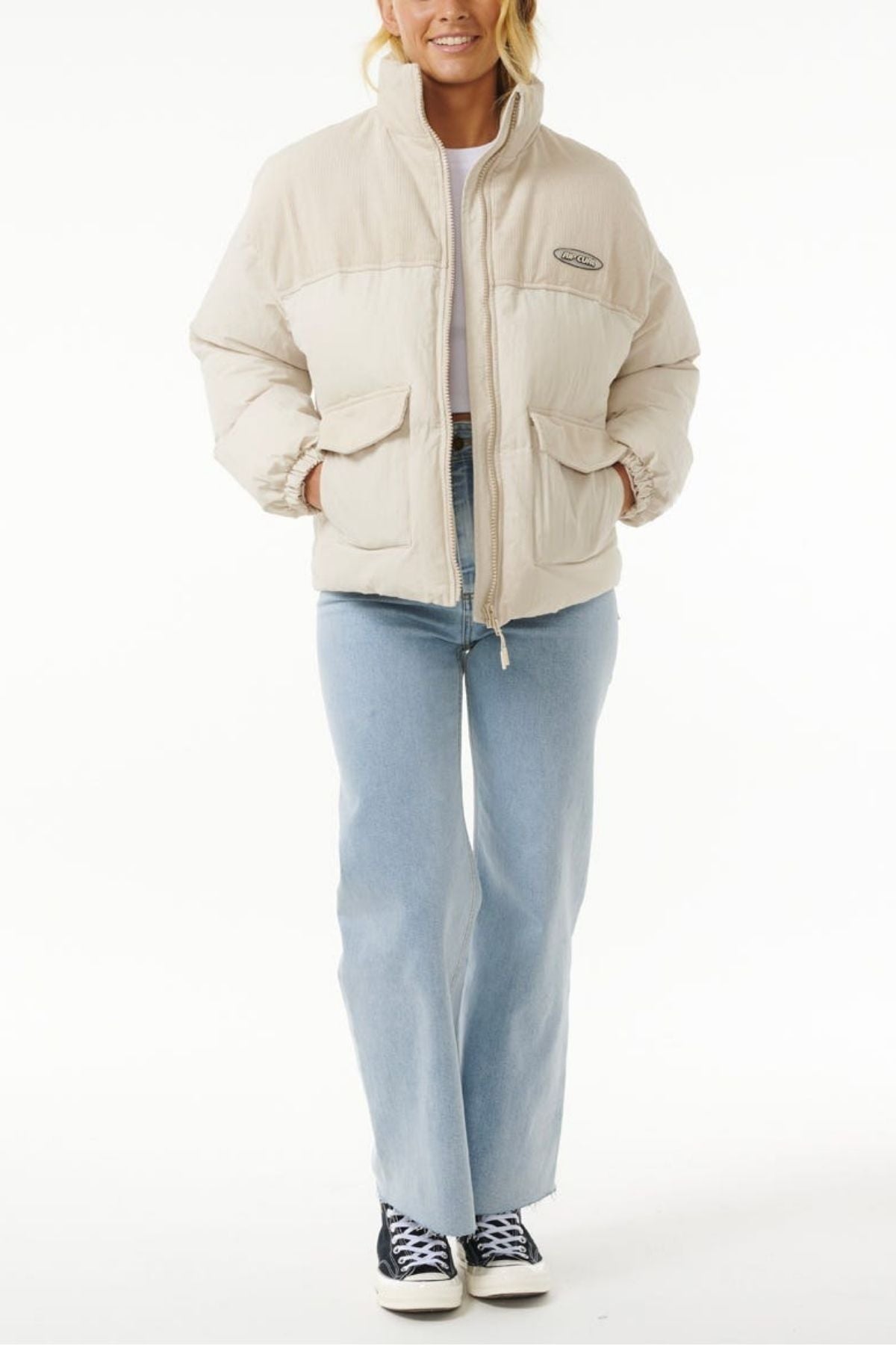 A person wearing the Rip Curl High Tide Mixed Cord Puffer, embroidered logo visible, light blue jeans, and black-and-white sneakers stands facing forward with hands in pockets against a plain white background.