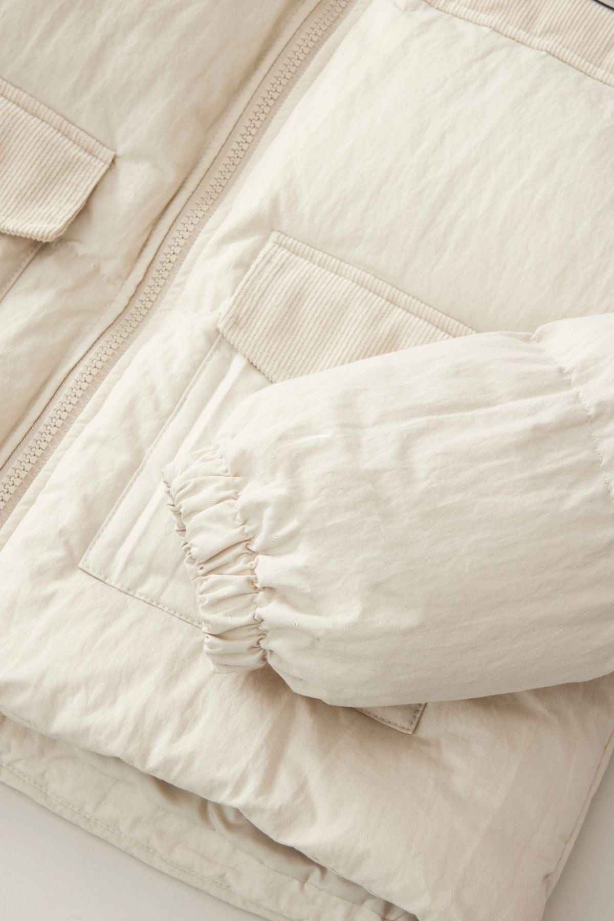 A close-up of the Rip Curl High Tide Mixed Cord Puffer in light beige, showing its zippered front, flap pockets, elasticated sleeve cuff, crinkled-texture fabric, and embroidered logo.