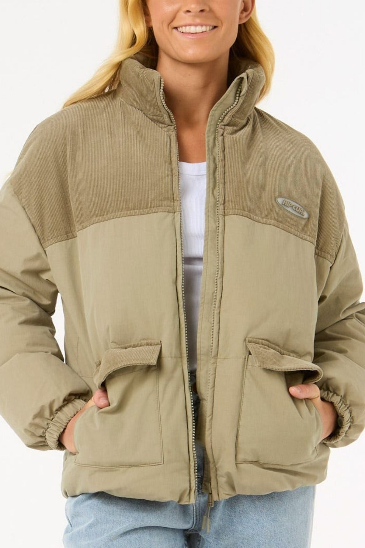 A woman smiles with hands in her pockets, wearing the Rip Curl High Tide Mixed Cord Puffer—a light brown jacket with corduroy accents and two front pockets—over a white top and light blue jeans.