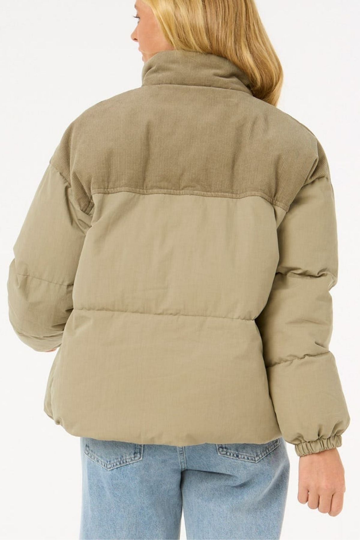 A woman with long blonde hair is seen from behind wearing the Rip Curl High Tide Mixed Cord Puffer with corduroy accents and an embroidered logo, paired with light blue jeans, set against a plain white background.