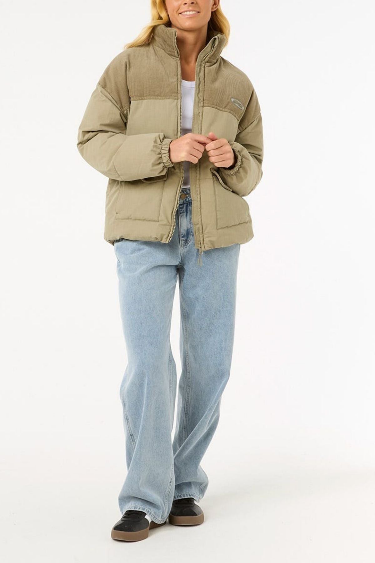 A person in a Rip Curl High Tide Mixed Cord Puffer jacket over a white top, blue wide-leg jeans, and black shoes stands against a plain white background.