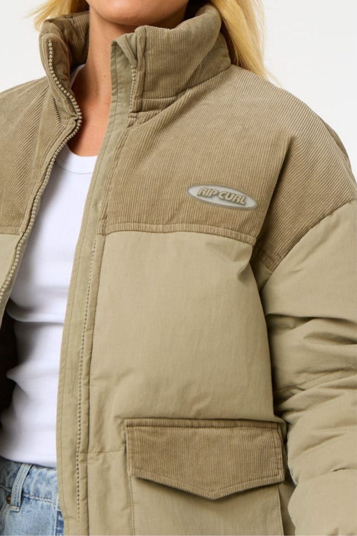 A person wears the Rip Curl High Tide Mixed Cord Puffer, a beige jacket with a high collar, embroidered logo on the chest, mixed fabric textures, large front pockets, and a white shirt underneath.