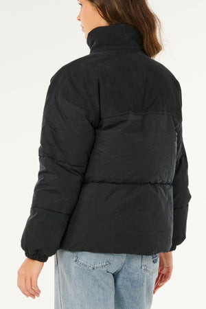 A person with long brown hair is seen from behind, wearing the Rip Curl High Tide Mixed Cord Puffer with a high collar and light blue jeans. The background is plain white.