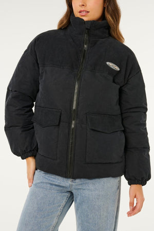 A woman stands against a plain white background wearing the Rip Curl High Tide Mixed Cord Puffer, a black jacket with large front pockets, high collar, visible front zipper, embroidered logo patch, and light blue jeans.