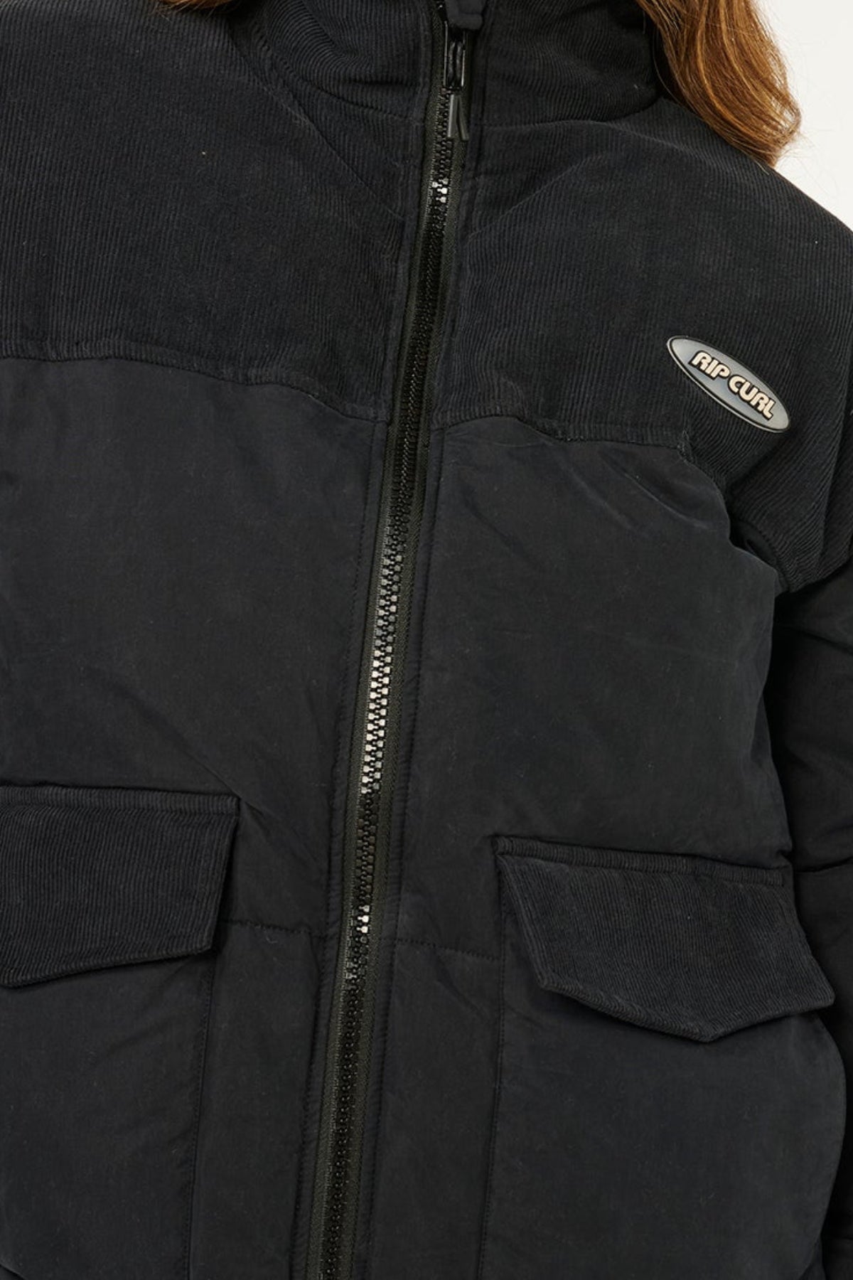 A close-up of the Rip Curl High Tide Mixed Cord Puffer, a black jacket with a central zipper, two front pockets, an embroidered logo patch on the left chest, and a corduroy-textured upper section.