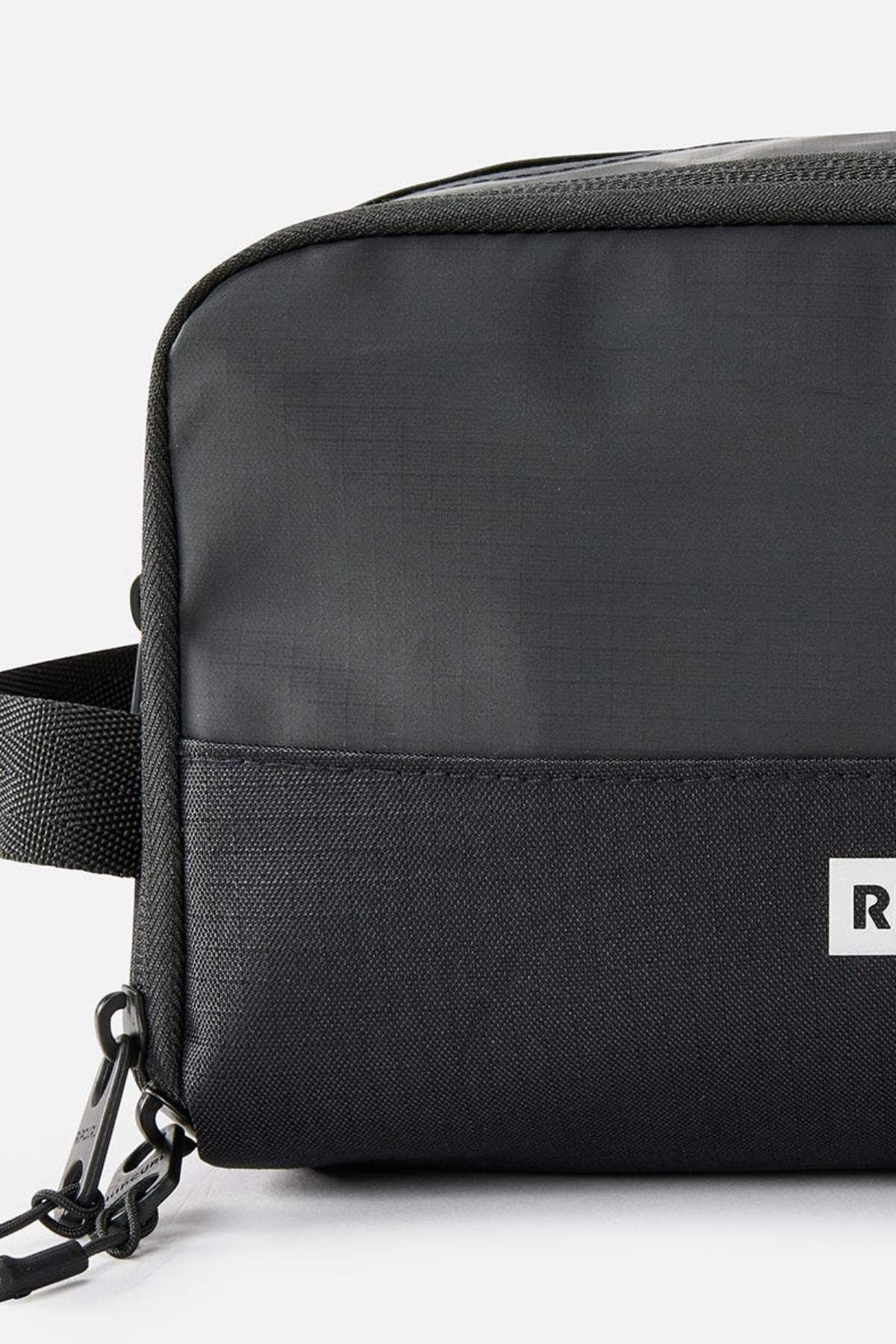 Close-up of the Rip Curl Icons Groom Toiletry Bag in a sleek black rectangular design, ideal for travel and organization. It boasts subtly textured fabric, a durable strap on the left, and a small white logo label on the lower right corner.