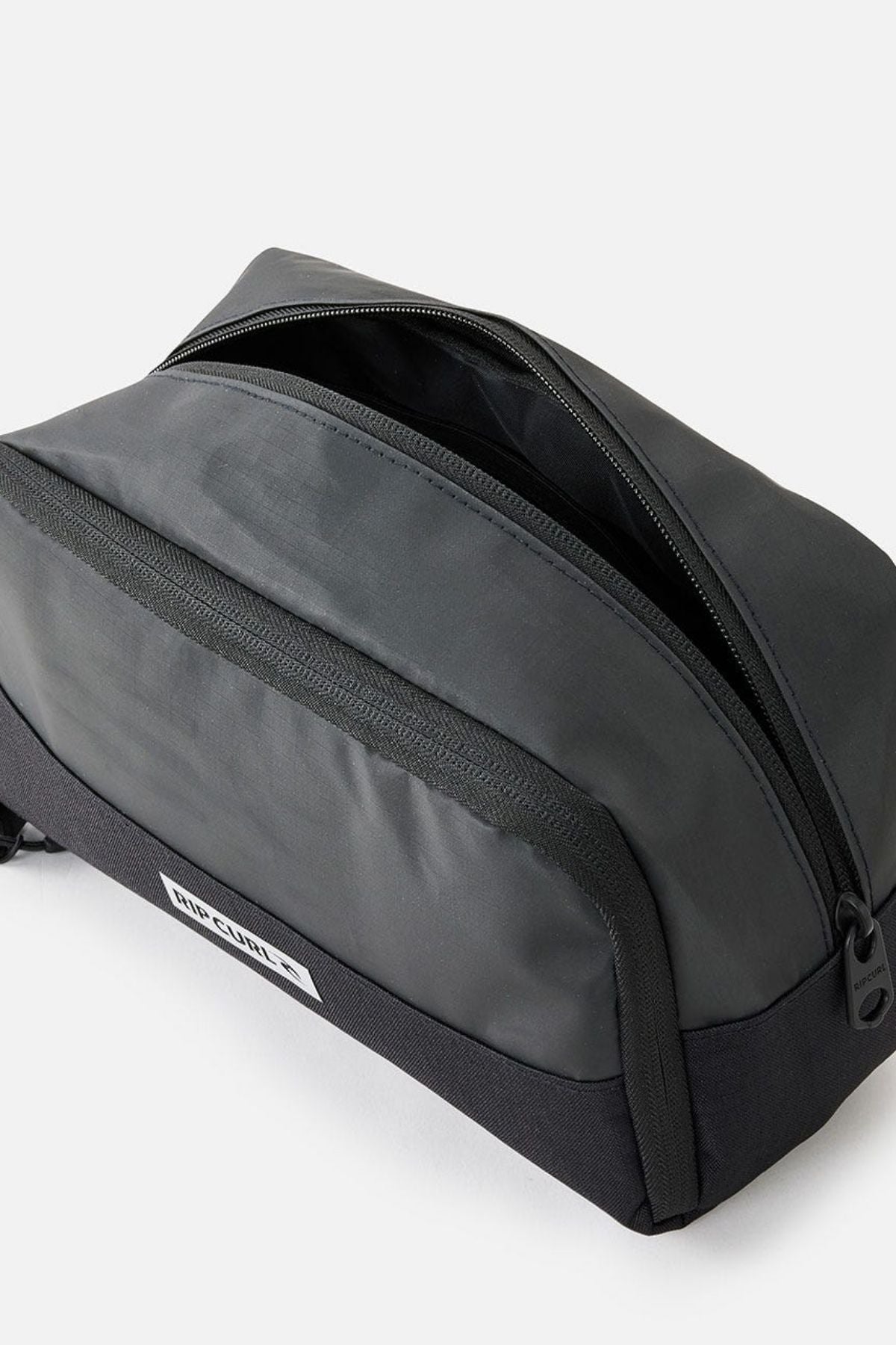 The Rip Curl Icons Groom Toiletry Bag, perfect for travel, comes in black and features a side pocket along with a small logo tag on the front. When opened, it reveals a spacious interior ideal for organization and is elegantly displayed on a plain, light-colored background.