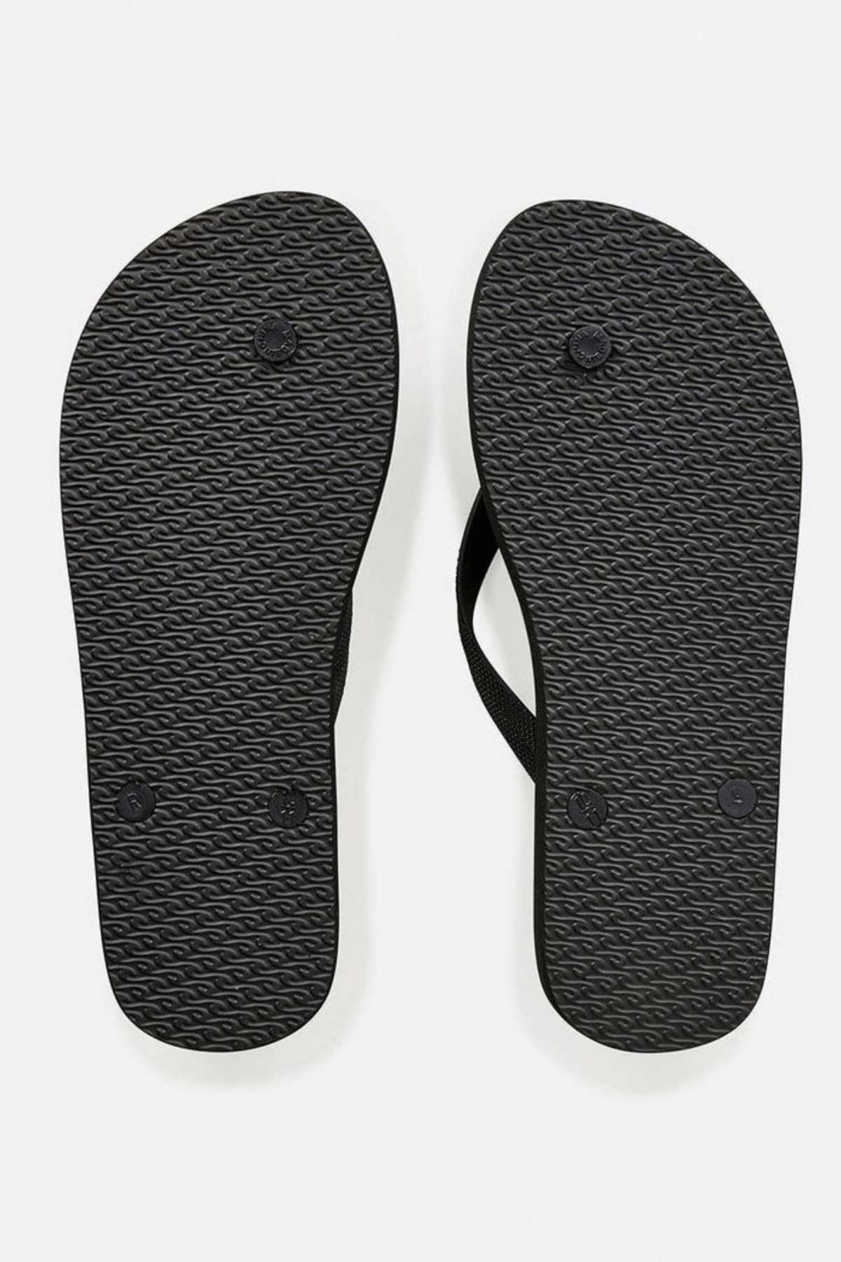 A pair of Rip Curl Icons of Surf Bloom Thongs is viewed from the bottom, highlighting their textured soles and ergonomic molded straps. Made with eco-friendly materials, these black flip-flops stand out against a plain white background.