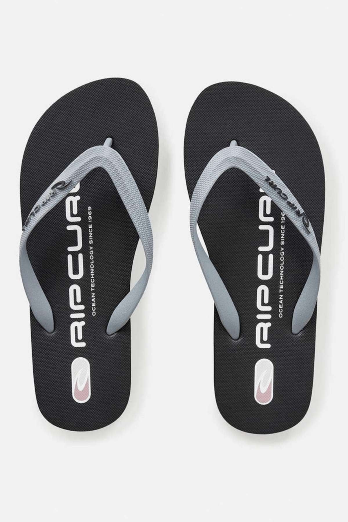 The Rip Curl Icons of Surf Bloom Thongs boast gray ergonomic molded straps set against black soles that prominently display the "Rip Curl" branding in white. Constructed with BLOOM™ algae blended foam EVA, these thongs embrace a minimalistic design that pays homage to the Rip Curl Icons of Surf heritage against a simple backdrop.