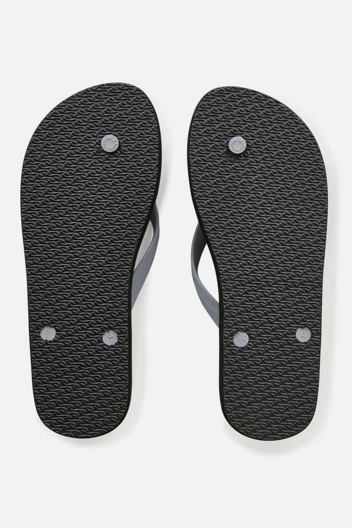 A pair of Rip Curl Icons of Surf Bloom Thongs in black is viewed from the bottom, showcasing textured soles made with BLOOM™ algae blended foam EVA and gray ergonomic molded straps. The image is set against a plain white background.