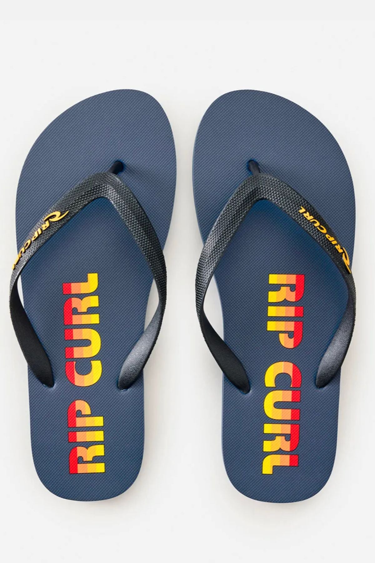 The Rip Curl Icons of Surf Bloom Thongs feature navy blue footbeds, black textured straps, bold red and yellow "RIP CURL" branding, and eco-friendly BLOOM algae-blended foam for comfort and sustainability.