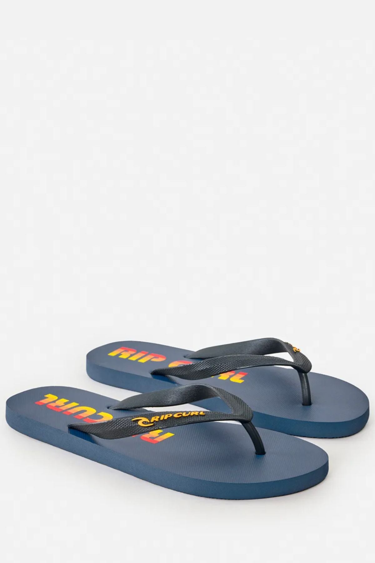 The Rip Curl Icons of Surf Bloom Thongs feature blue footbeds with dark straps and a yellow, red, and orange logo. Made with eco-friendly BLOOM algae-blended foam, they're shown on a light grey background.