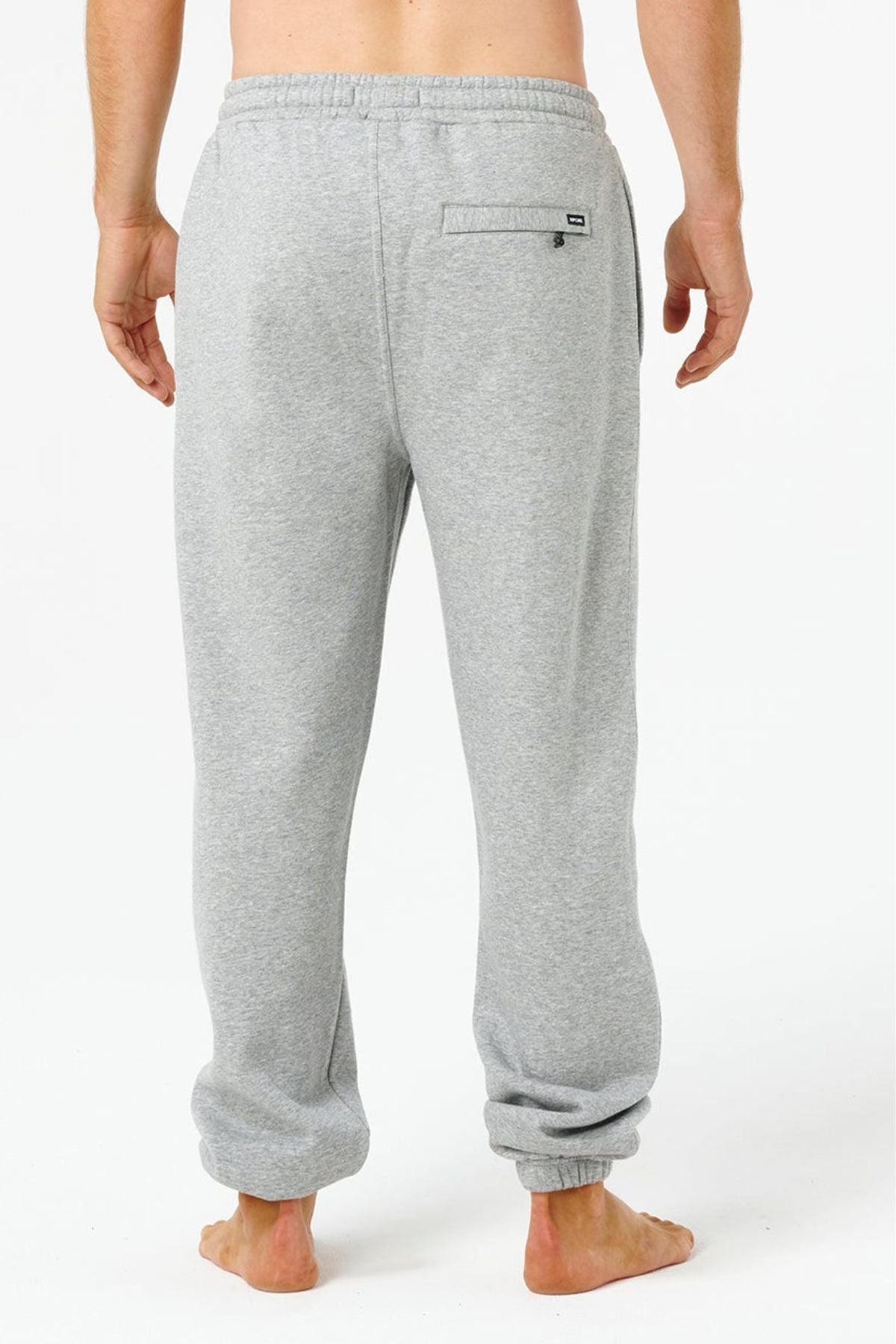 A person stands barefoot, facing away, wearing Rip Curl Icons Of Surf Trackpant in light gray with an elastic waistband and a single back pocket, set against a plain white background.